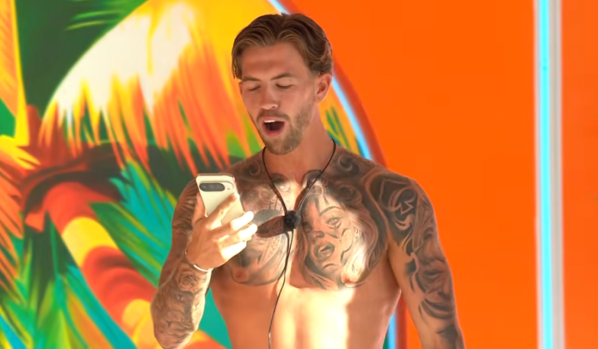 A shirtless, tattooed man with an open mouth reads from a smartphone.