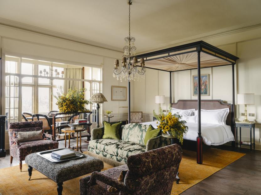 A luxurious hotel room with a four-poster bed, patterned sofa and chairs, and a large window overlooking a garden.