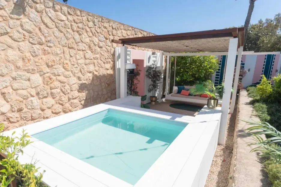 A patio with a pool in the foreground, a stone wall on the left, and a covered swing couch in the background.