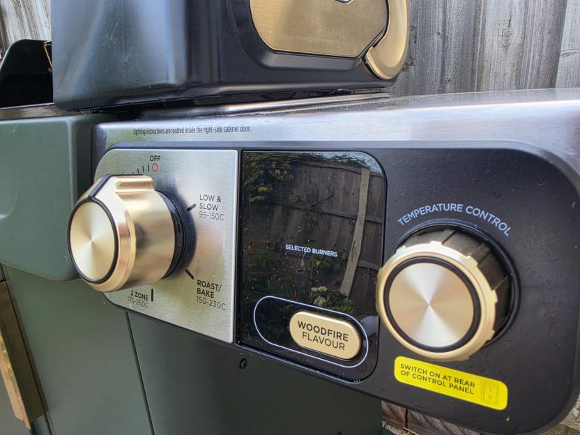Close-up of a grill's control panel, featuring golden temperature knobs, an LED display, and settings for "Low & Slow," "Roast/Bake," "2 Zone," and "Woodfire Flavour."