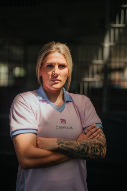 Jen Beattie, an Arsenal football player and breast cancer advocate, stands with her arms crossed, wearing a pink and blue polo shirt with "Breast Cancer Now" and "Novartis" logos.