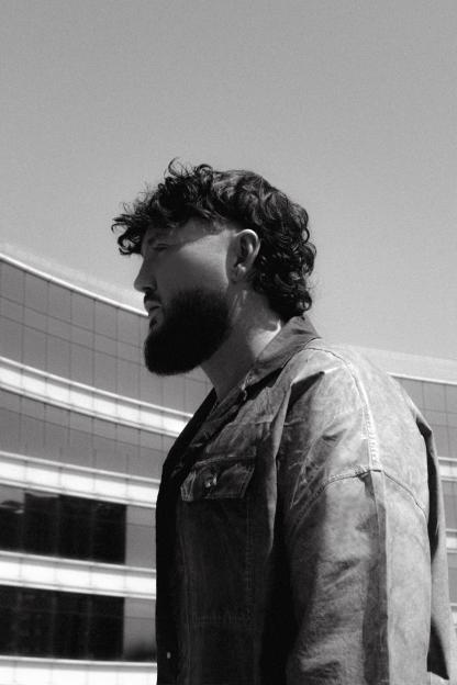 Black and white profile of singer James Arthur looking right, with a large, dark beard and curly hair, wearing a dark jacket, against a building.