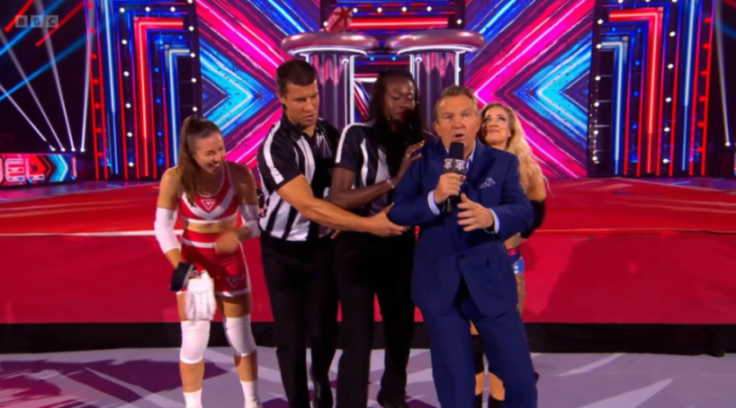 Two referees escorting TV host Bradley Walsh away while other Gladiators competitors laugh.