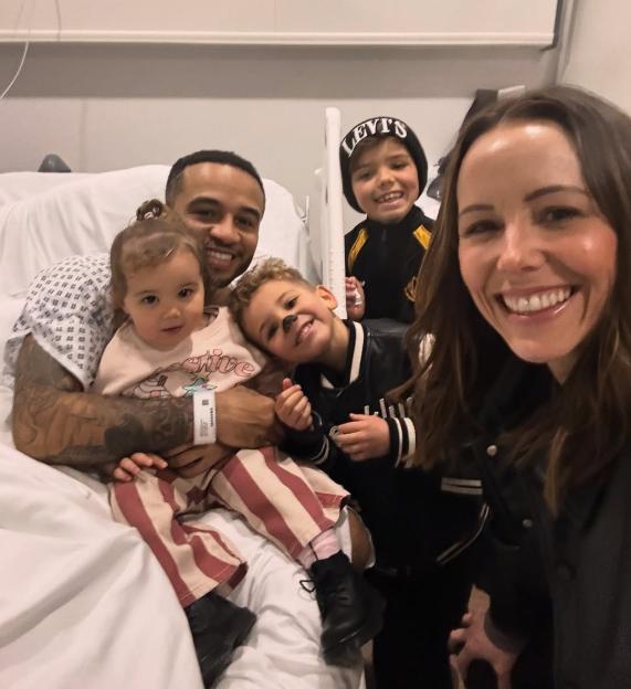 A family of five, including a father in a hospital bed, mother, and three children, smiles for a selfie.