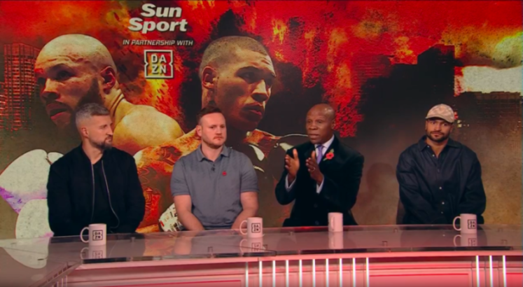 Four men sit at a table on a stage in front of a promotional boxing poster.