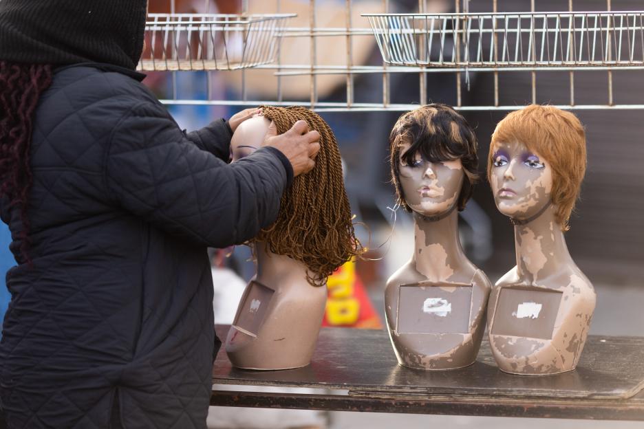 A person placing a braided wig onto a mannequin head at a market stall.