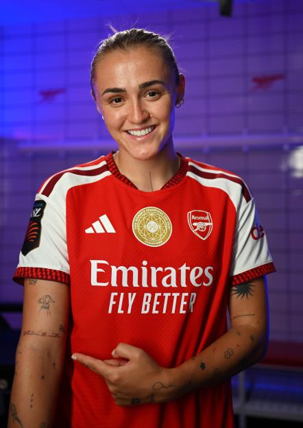 Georgia Stanway smiling and pointing to a tattoo on her arm, wearing an Arsenal FC jersey.