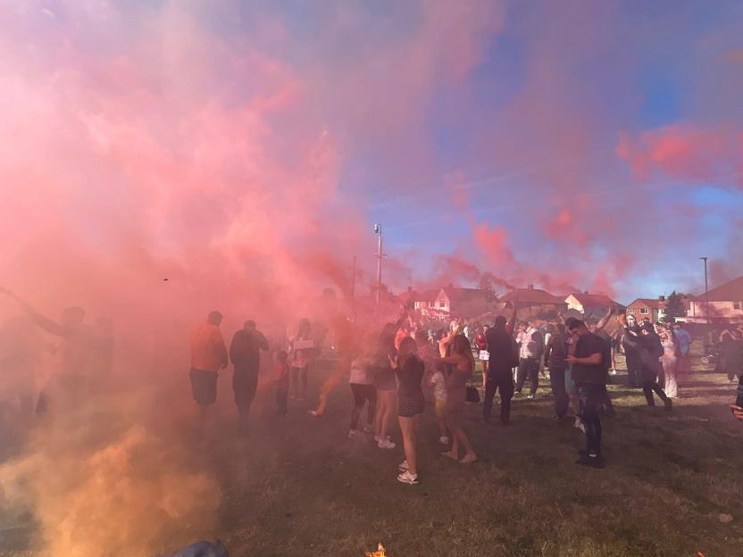A crowd of people on a grassy field enveloped in bright pink and orange smoke from flares.