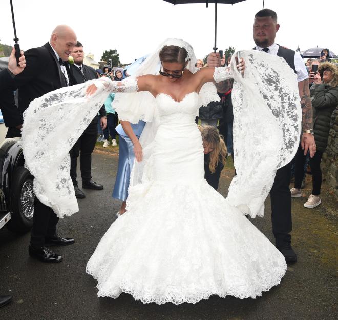 Venezuela Fury in a wedding dress, being helped by Tyson Fury and another man, under an umbrella.