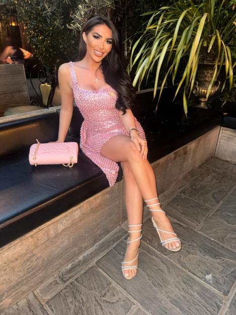 A woman in a pink sequin dress and spiral-strap heels sits on a bench.