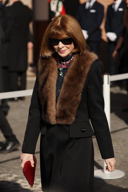 Anna Wintour in a black coat with a brown fur collar and a red clutch.