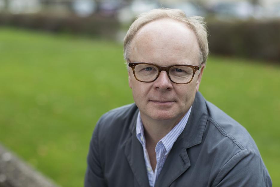 Actor Jason Watkins, wearing glasses and a gray jacket over a striped shirt, looking directly at the camera at the Cheltenham Literature Festival.