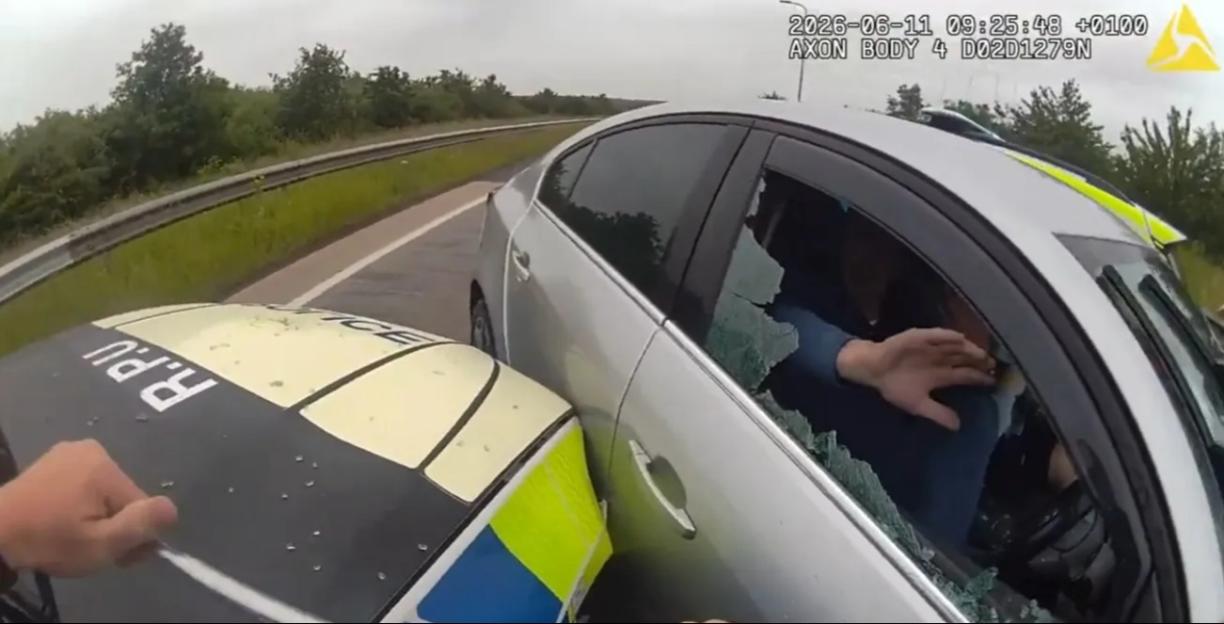 A police officer looking into the smashed window of a silver car on the side of a highway.