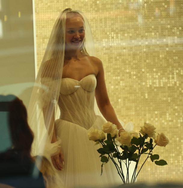 Roxy Horner in a white strapless wedding gown and veil, smiling with a bouquet of light-colored roses in front of a golden sparkling background.