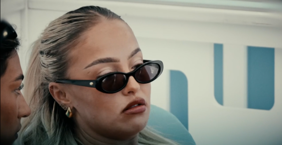 A woman wearing dark oval sunglasses and light hair is looking intently to the right, with another person's profile visible on the left.
