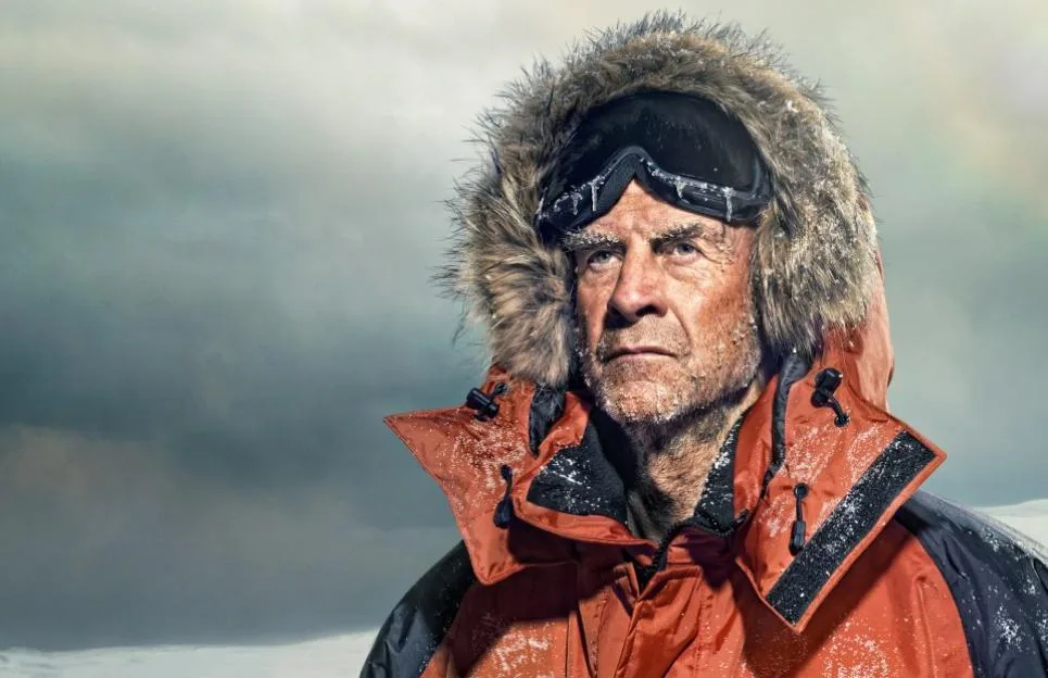 Sir Ranulph Fiennes, explorer, in an orange parka with a fur-trimmed hood and goggles, with snow and ice on his face and clothing.