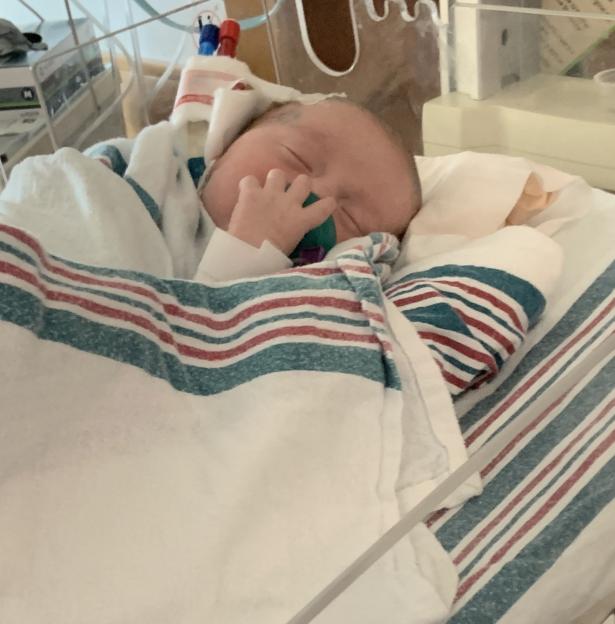 Newborn Leo Debelak in a hospital bed with a pacifier, wrapped in a striped blanket.