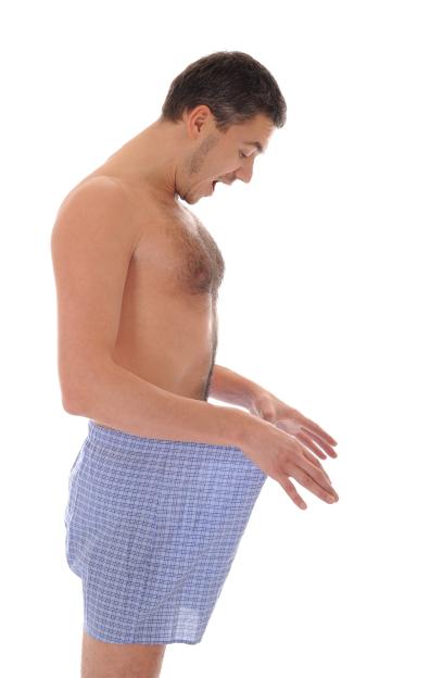 A shirtless man in blue plaid boxers looking down into his pants with a surprised expression.