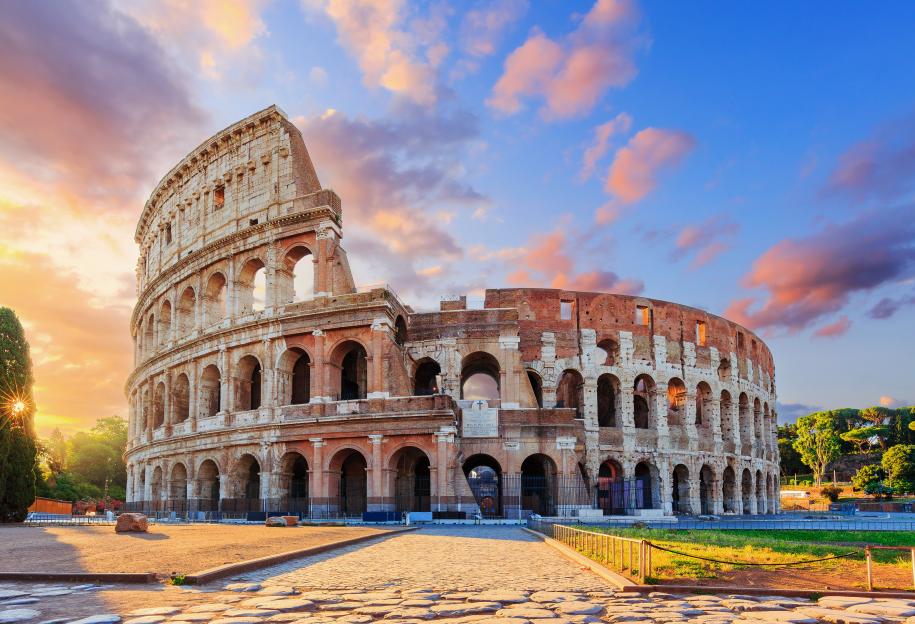 The Colosseum at sunrise, with warm sunlight illuminating its ancient stone arches against a vibrant sky of blue and soft pink clouds.