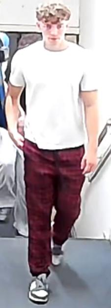 A young man with light, curly hair, wearing a white t-shirt, maroon patterned trousers, and white and black athletic shoes.