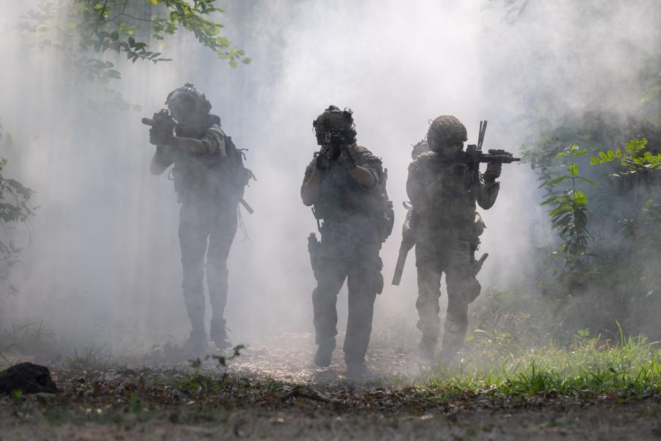 Three armed soldiers in military uniforms walking through heavy smoke in a forest.