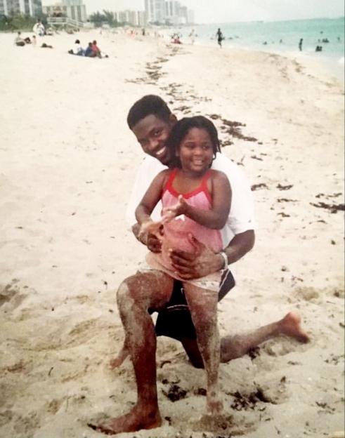 A smiling man on one knee holding a young girl in a pink swimsuit, both covered in sand on a beach.