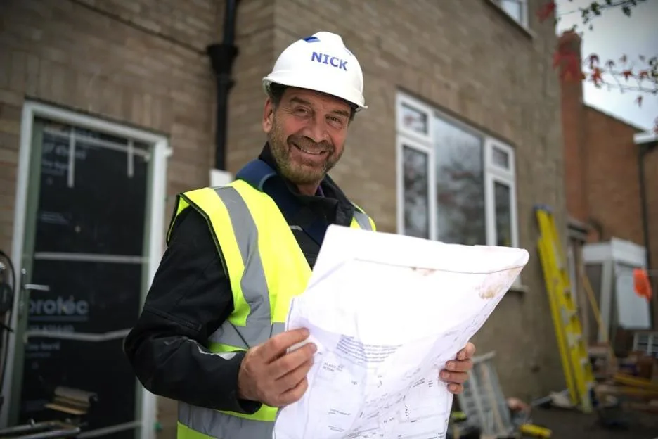 A smiling construction worker wearing a white hard hat with the name "Nick" and a high-visibility yellow vest, holds up building plans in front of a house under construction.