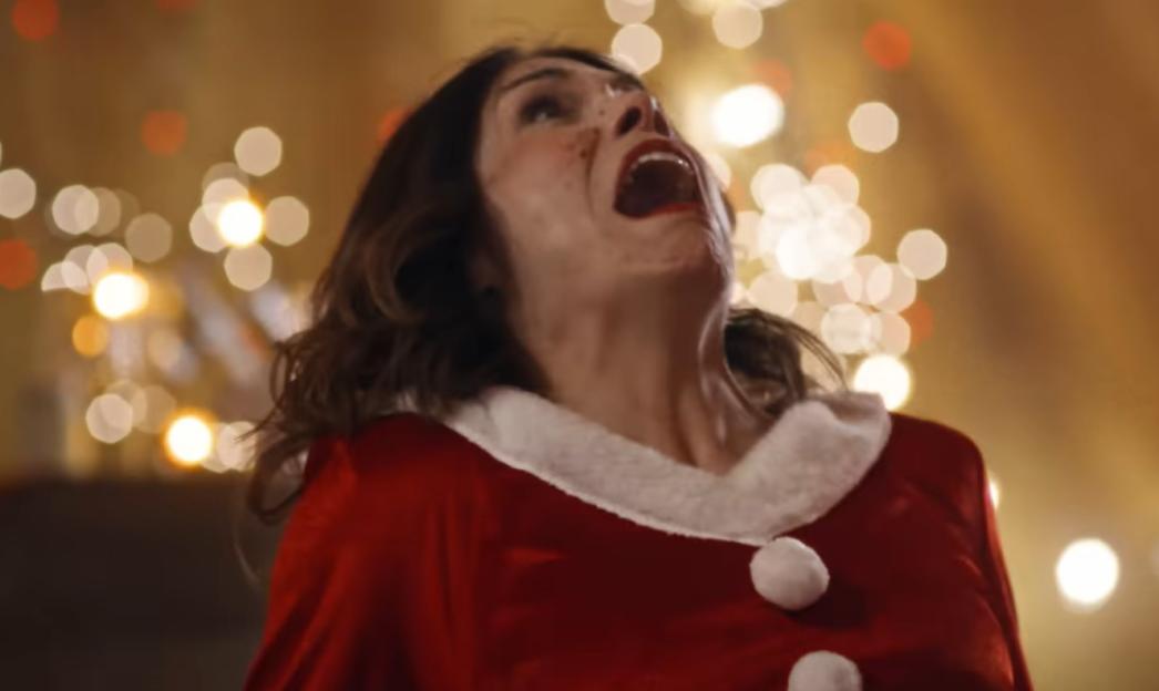 A woman in a red and white Santa costume with a terrified, open-mouthed expression looks upwards with blurred Christmas lights in the background.