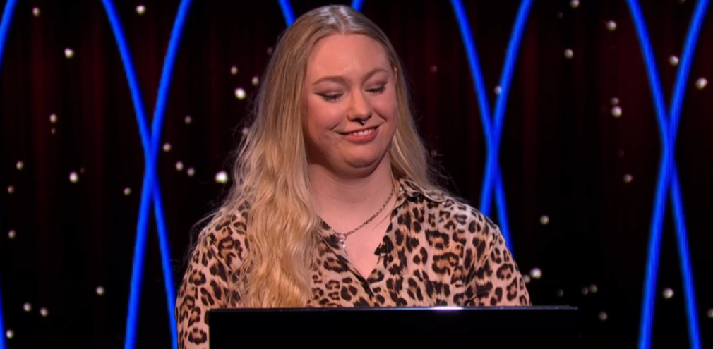 A young woman with long blonde hair and a leopard print shirt smiles, seated in front of a dark background with blue glowing lines and sparkling lights.