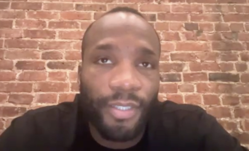 A man speaking in front of a brick wall.
