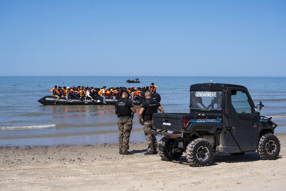 Two gendarmerie officers and a Polaris Ranger ATV on a beach, watching an inflatable boat full of migrants wearing orange life vests in the water.