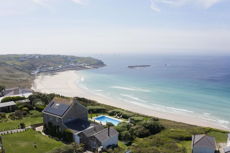 An aerial view of a stone house with solar panels on a cliff overlooking a beach, a town, and the Atlantic Ocean.