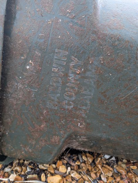 Close-up of a weathered green wheelie bin with "BALDWIN COUNTY, ALABAMA" embossed on its side, surrounded by barnacles and resting on pebbles.