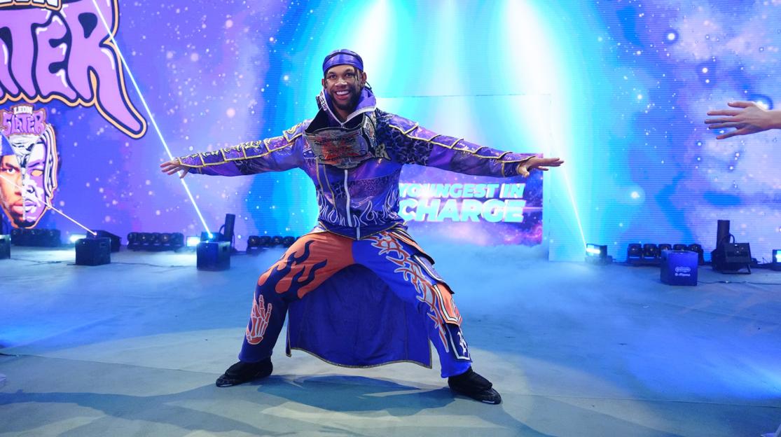 A wrestler wearing a purple and orange outfit with a championship belt, arms extended, on a stage with a starry blue background.