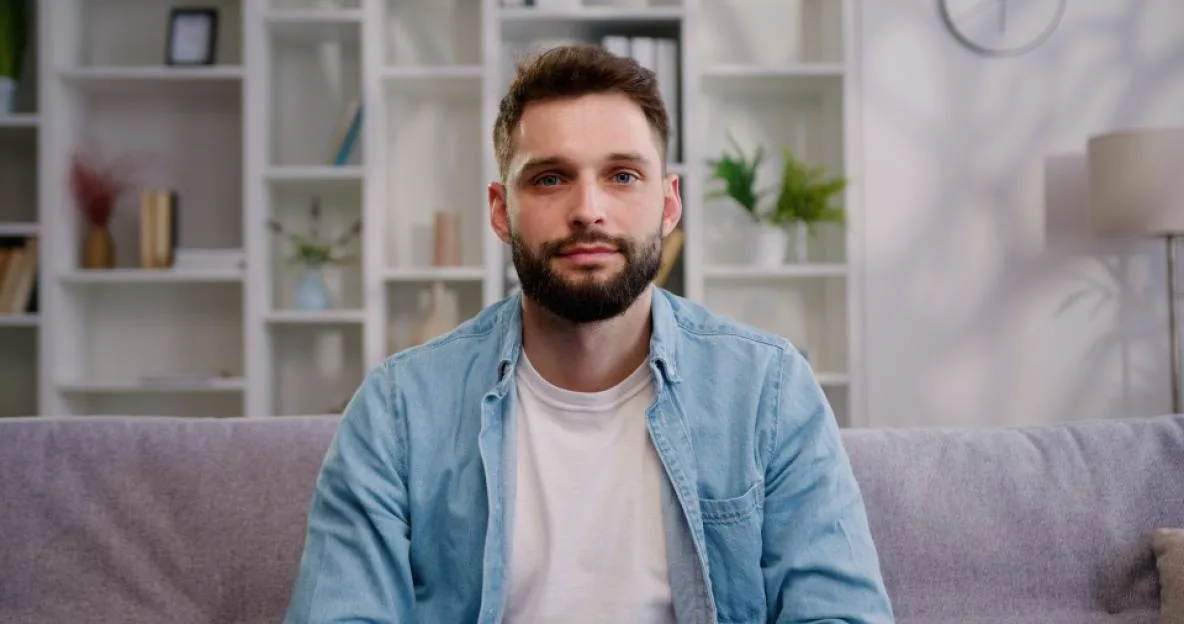 Portrait of a bearded man in a denim shirt sitting on a couch.