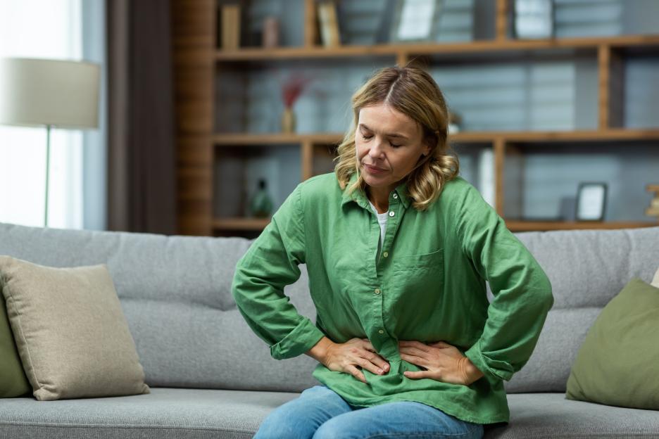 An elderly woman is sitting on the sofa at home, holding her stomach with her hands. Feels the pain of internal organs, indigestion, poisoning, menstrual pain