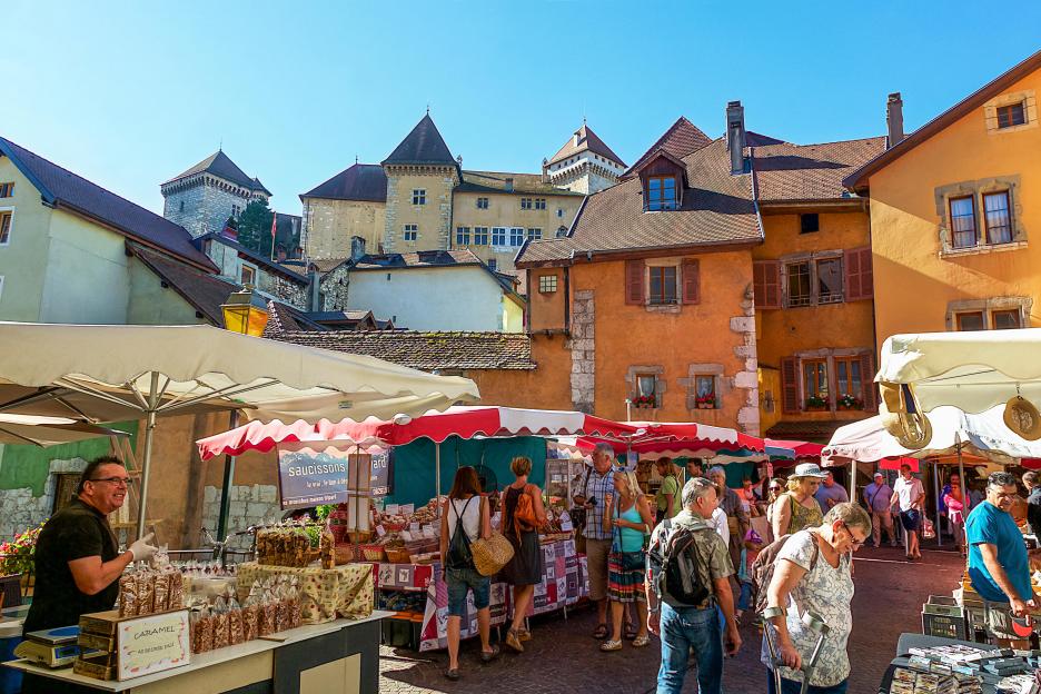 People browse goods at the outdoor markets of Annecy, France, with old buildings and a tower in the background.