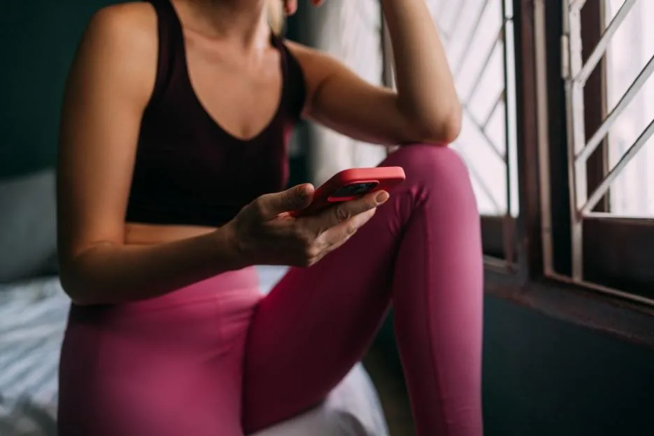 An Unrecognizable Beautiful Female In Sports Clothes Sitting On Her Bed Near The Window And Watching Something On Her Red Smartphone After Her Daily Workout