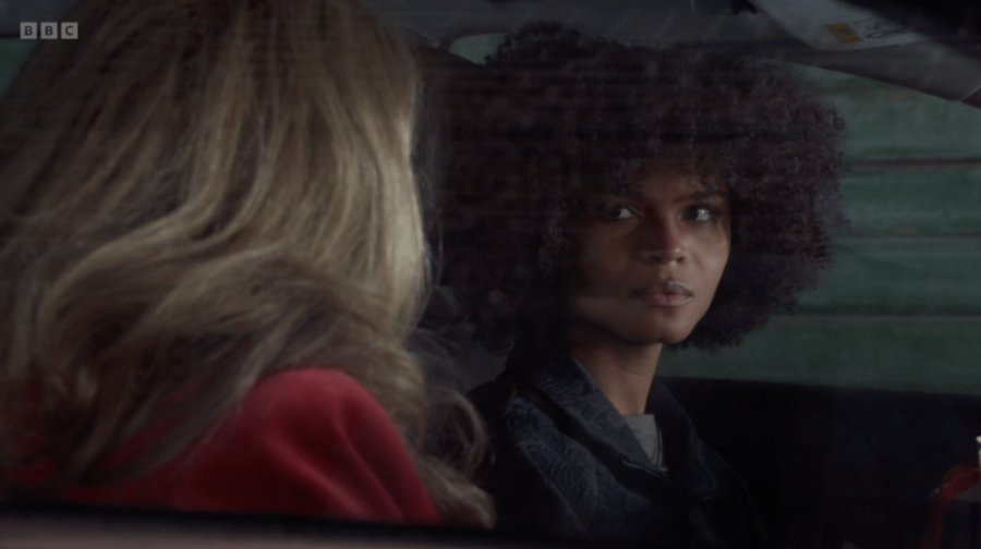 A woman with dark, curly hair looks over her shoulder at a blonde woman in a red top.