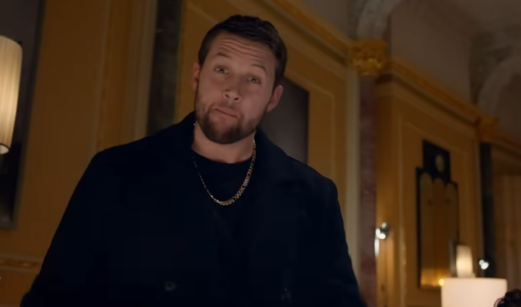A man with a beard and gold chain looks down at the camera.
