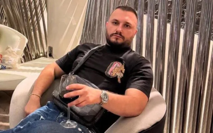 A man with a beard and short hair sitting in a chair, holding a glass of red wine.