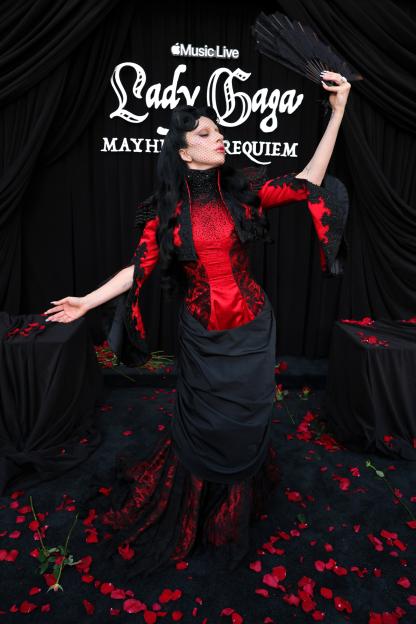 Lady Gaga in a dramatic red and black gothic outfit with a fan, posing on a black carpet covered in red rose petals, in front of a "Lady Gaga MAYHEM REQUIEM" sign.