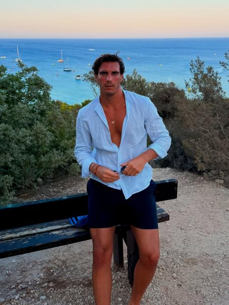 Lorenzo Alessi in a light blue shirt and dark shorts standing by a wooden bench with the sea and sailboats in the background.