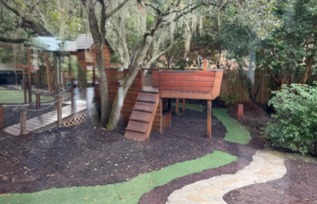 A playground with a wooden pirate ship structure, rope bridge, and pathways of artificial turf and stone.