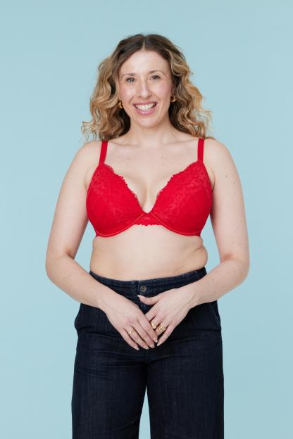 A smiling woman with blonde curly hair, wearing a red lace bra and dark wash jeans, stands against a light blue background.