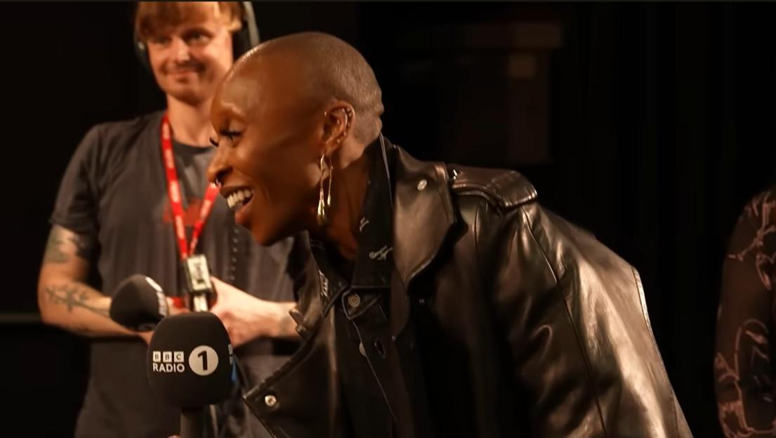 A person wearing a leather jacket and gold earrings, smiling at a BBC Radio 1 microphone.