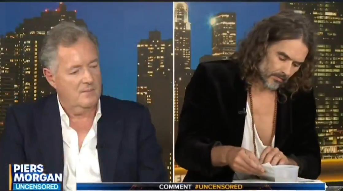 Piers Morgan and Russell Brand sitting at a table with city skylines in the background.
