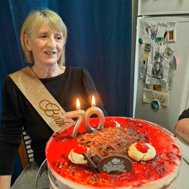 A woman wearing a "Birthday" sash sits next to a birthday cake with 70-shaped candles.