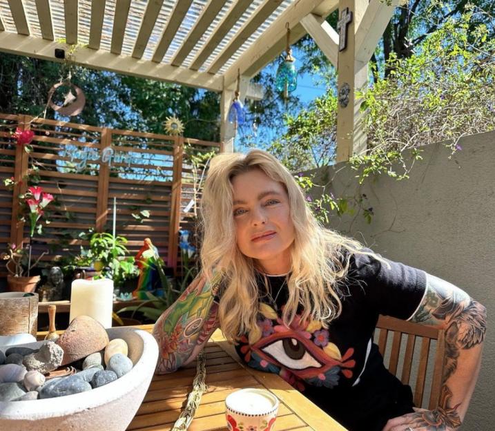 Erika Eleniak posing with tattoos on her arms, seated outdoors at a wooden table with a bowl of rocks and a candle.