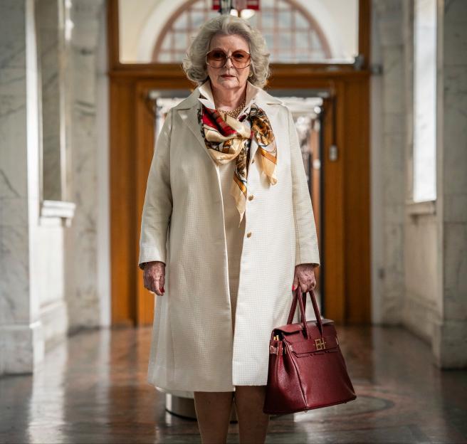 Brenda Blethyn as Emma Harte in 1970s fashion, wearing a white coat, a patterned scarf, and carrying a red handbag.